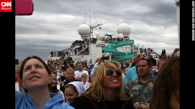 Passengers fill the deck of the ship and look up toward the sky. 